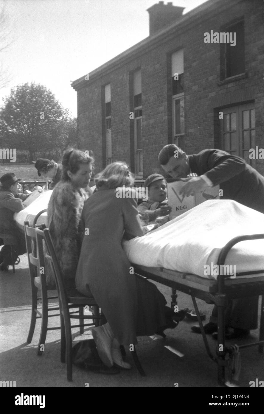 1942, historical, May, wartime and a young boy scout with parents beside his hospital bed outside in the courtyard for the fresh air at the Childrens Hospital, Carlshalton, Surrey, England, UK. On land owned by the Metropolitan Asylums Board, a hospital was built in 1908, the Southern Hospital. The following year the MAB took over responsibilty for 'sick or convalescent or debilitated children' and it became the Childrens Hospital, renamed as the Queen Mary's Hospital for Children in 1914. Care was provided in 24 blocks of four U-shaped cottages. It closed in 1993. Stock Photo