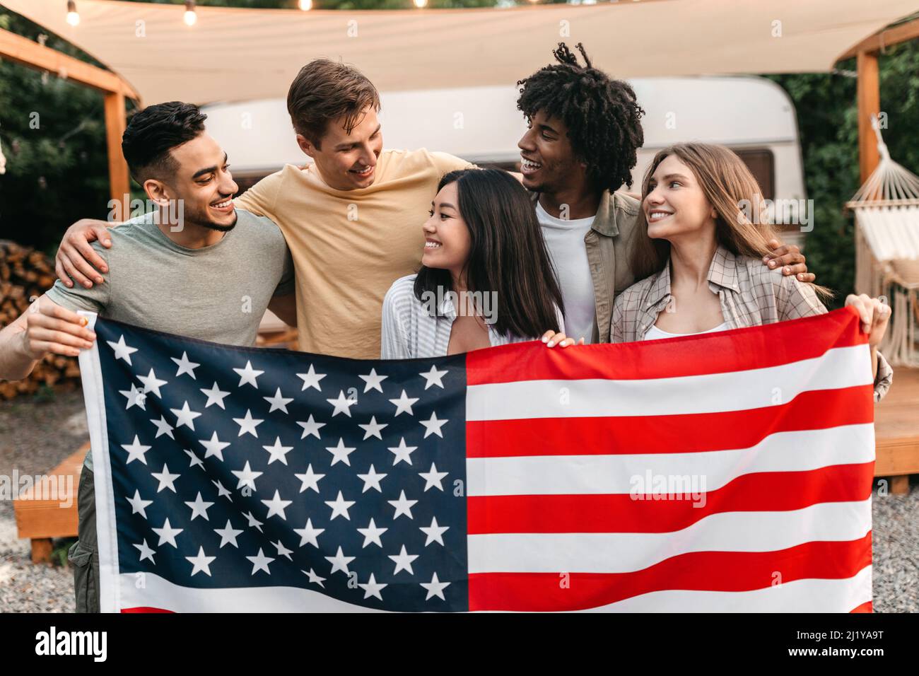 Millennial interracial friends holding American flag and smiling in front of RV at campsite Stock Photo