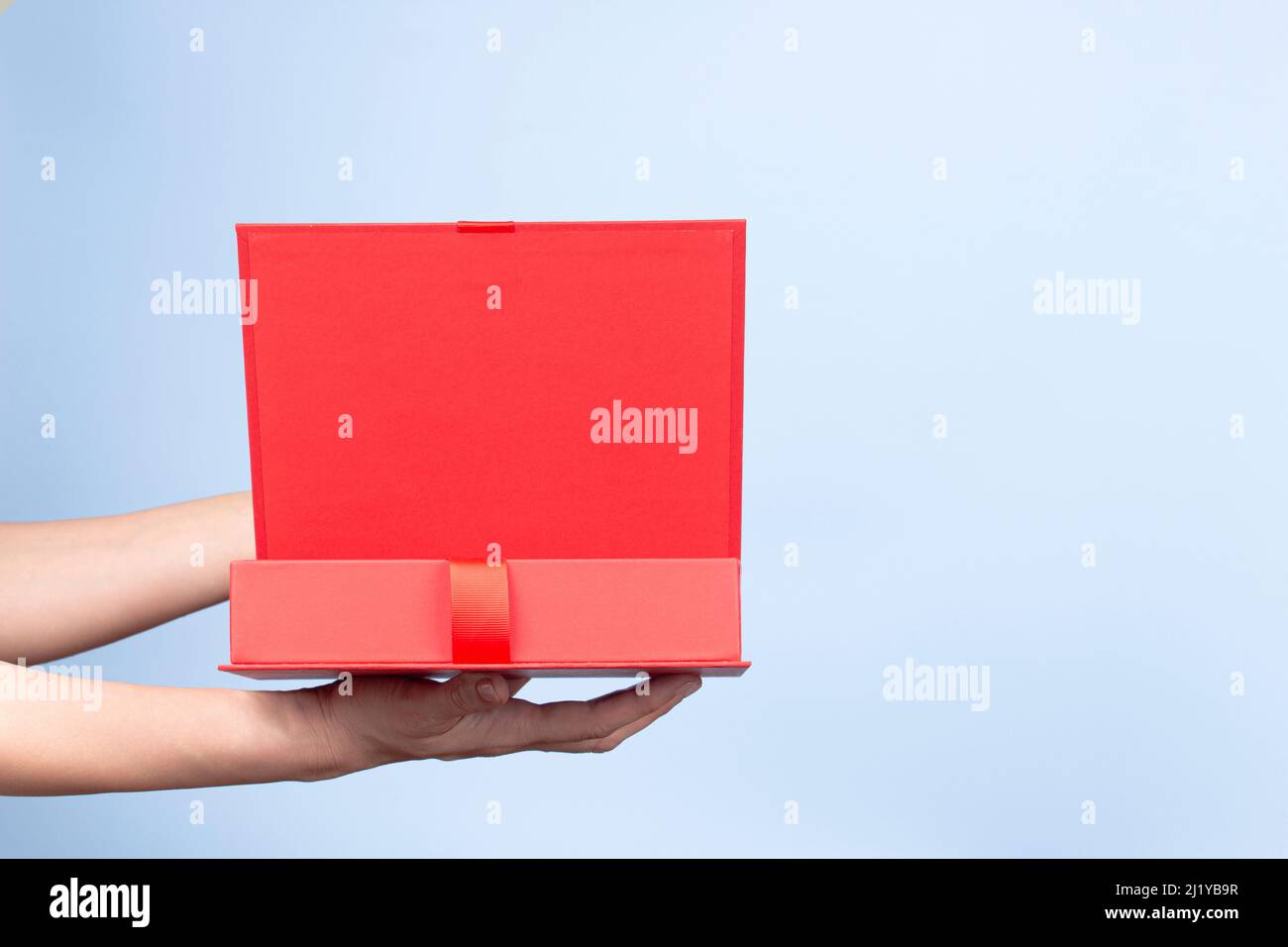Female hands opening red elegant luxury box with ribbon. Mockup present gift box on light blue background. Front view Stock Photo