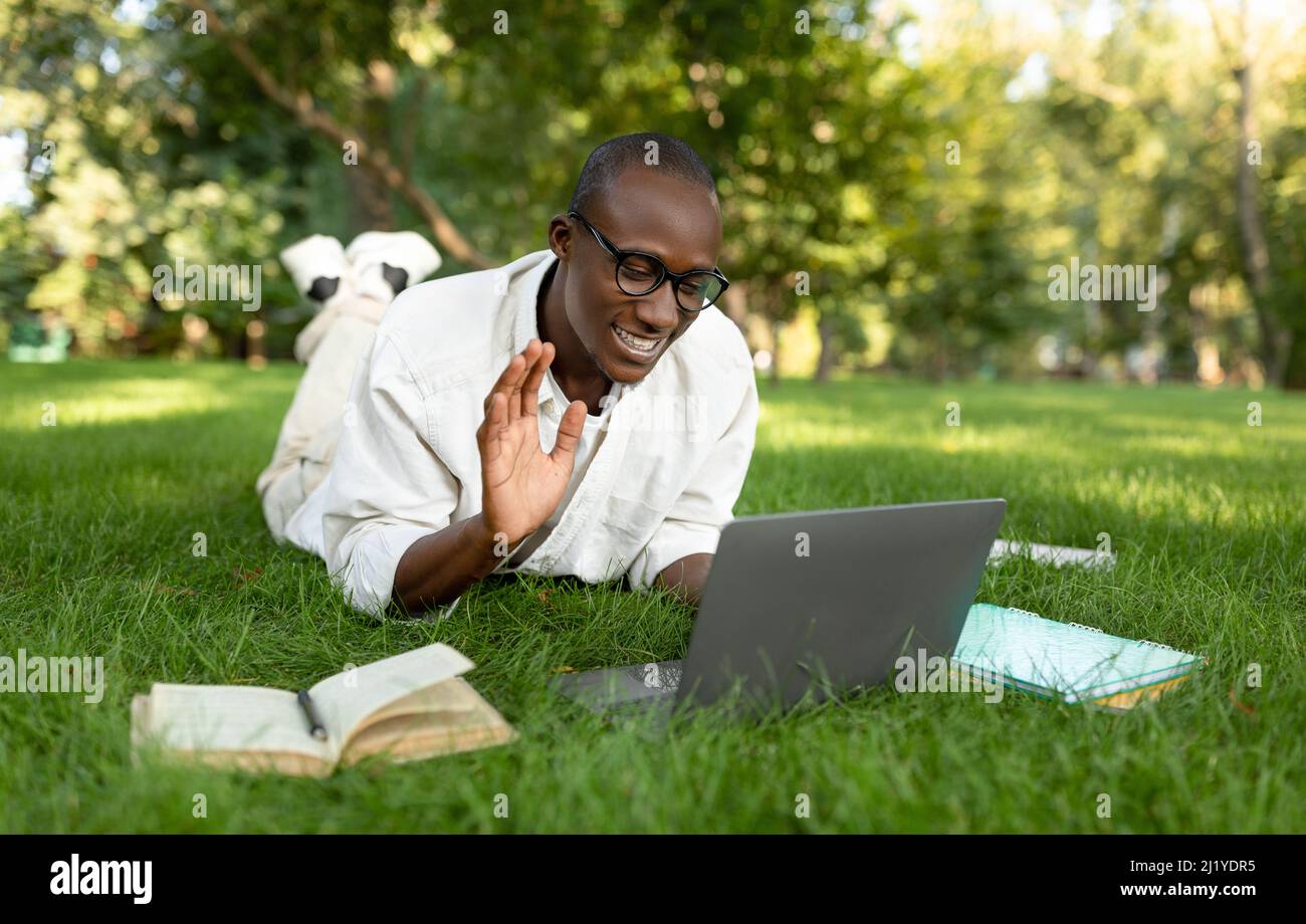 Remote education concept. Black male student video calling via laptop, having online lecture, waving at webcam outdoors Stock Photo