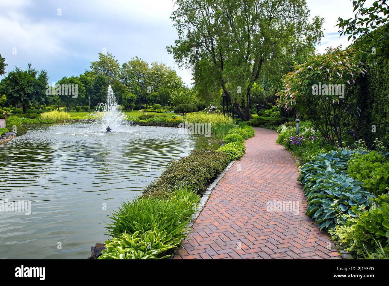 A pond filled with water with a spray jet fountain in a park with pedestrian sidewalks made of stone tiles among different plants, landscape design of Stock Photo