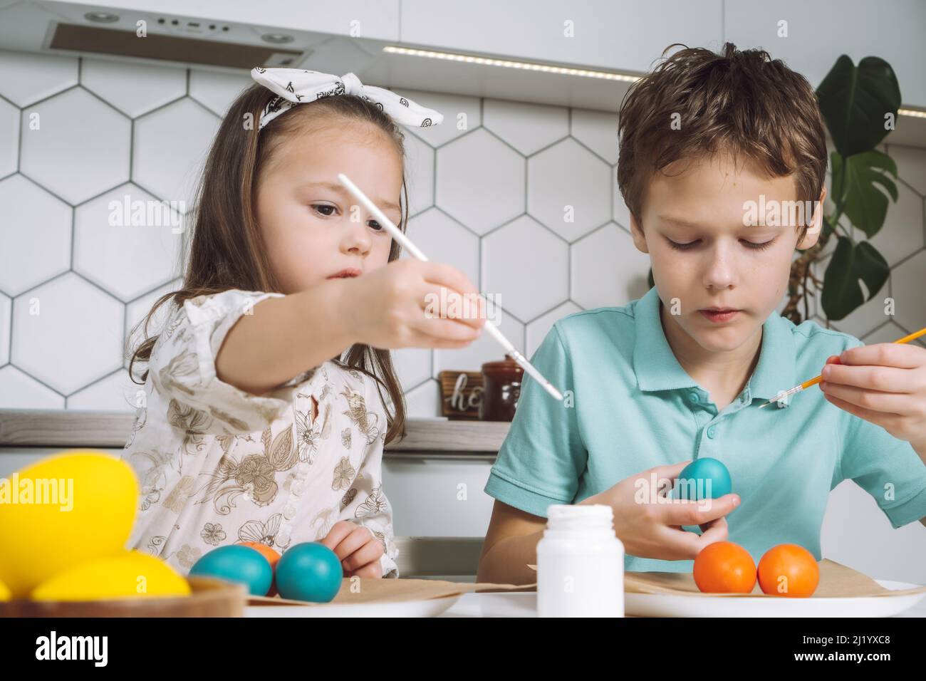 Portrait of enthusiastic two little children, boy and girl, painting brush multi coloring Easter hens eggs paper plate table, plant in kitchen. Making Stock Photo
