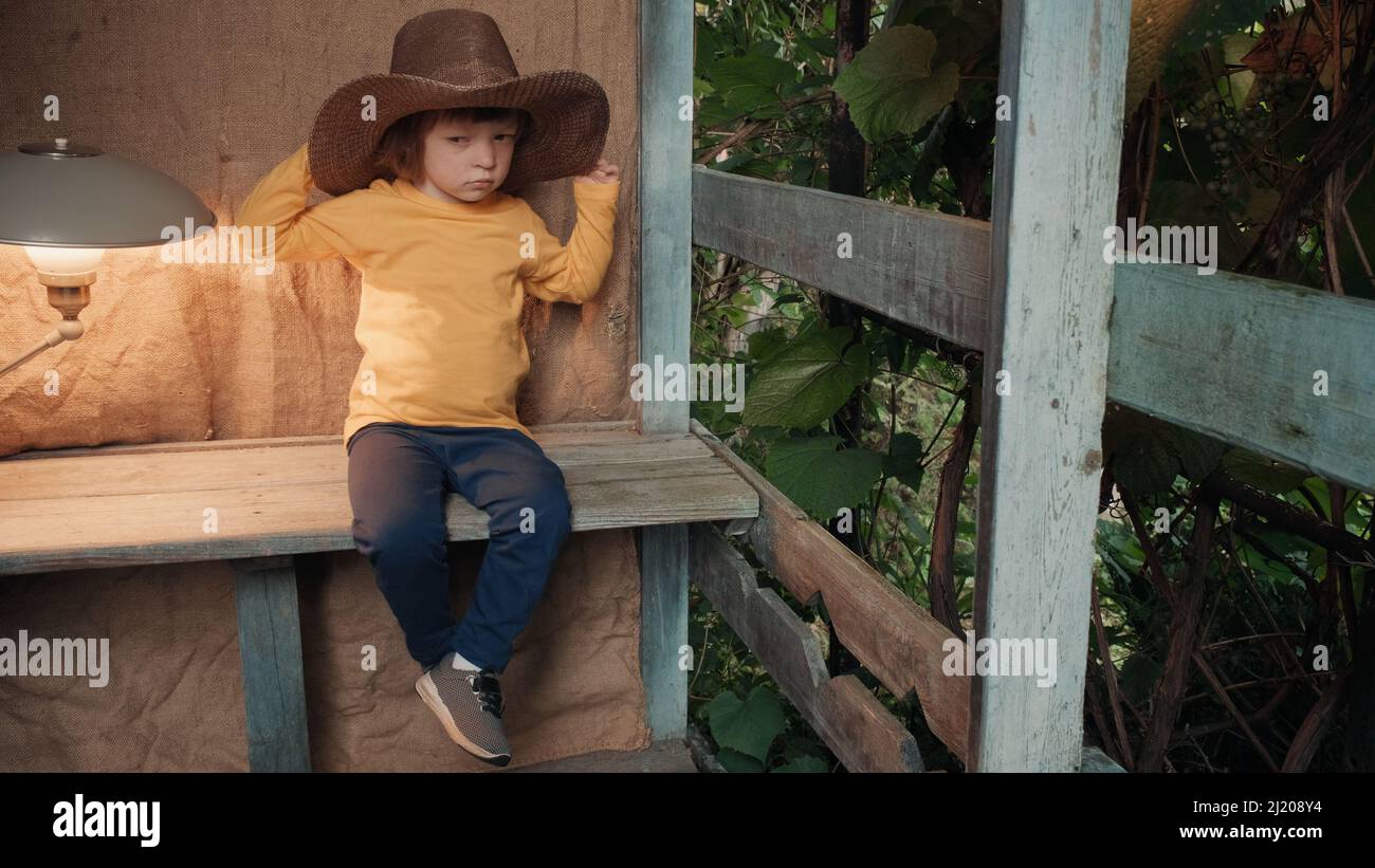 Small, funny child in big cowboy hat is sitting on wooden porch of country house Stock Photo