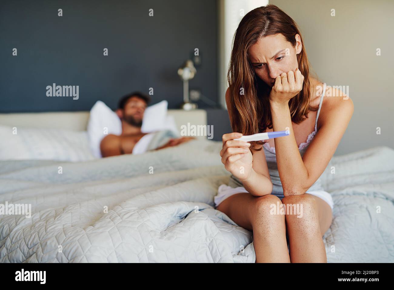 Please be negative.... Shot of a woman looking worried while holding a pregnancy test with her partner lying in the background. Stock Photo