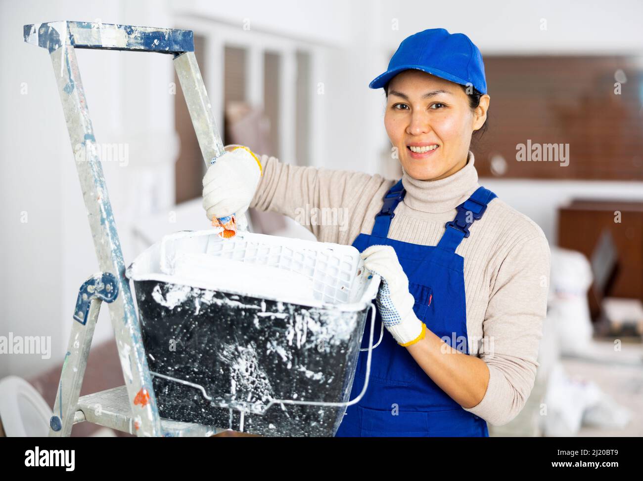 Woman painter in overalls with paint roller in building under ...