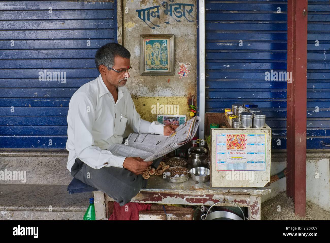 A middle-aged Indian betel (paan) vendor sits at his paan stall reading a newspaper; Mumbai, Maharashtra, India Stock Photo