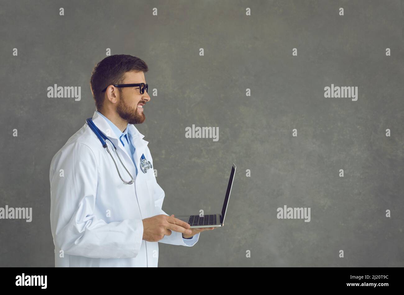 Smiling doctor in white coat working using laptop side view shot on grey wall Stock Photo