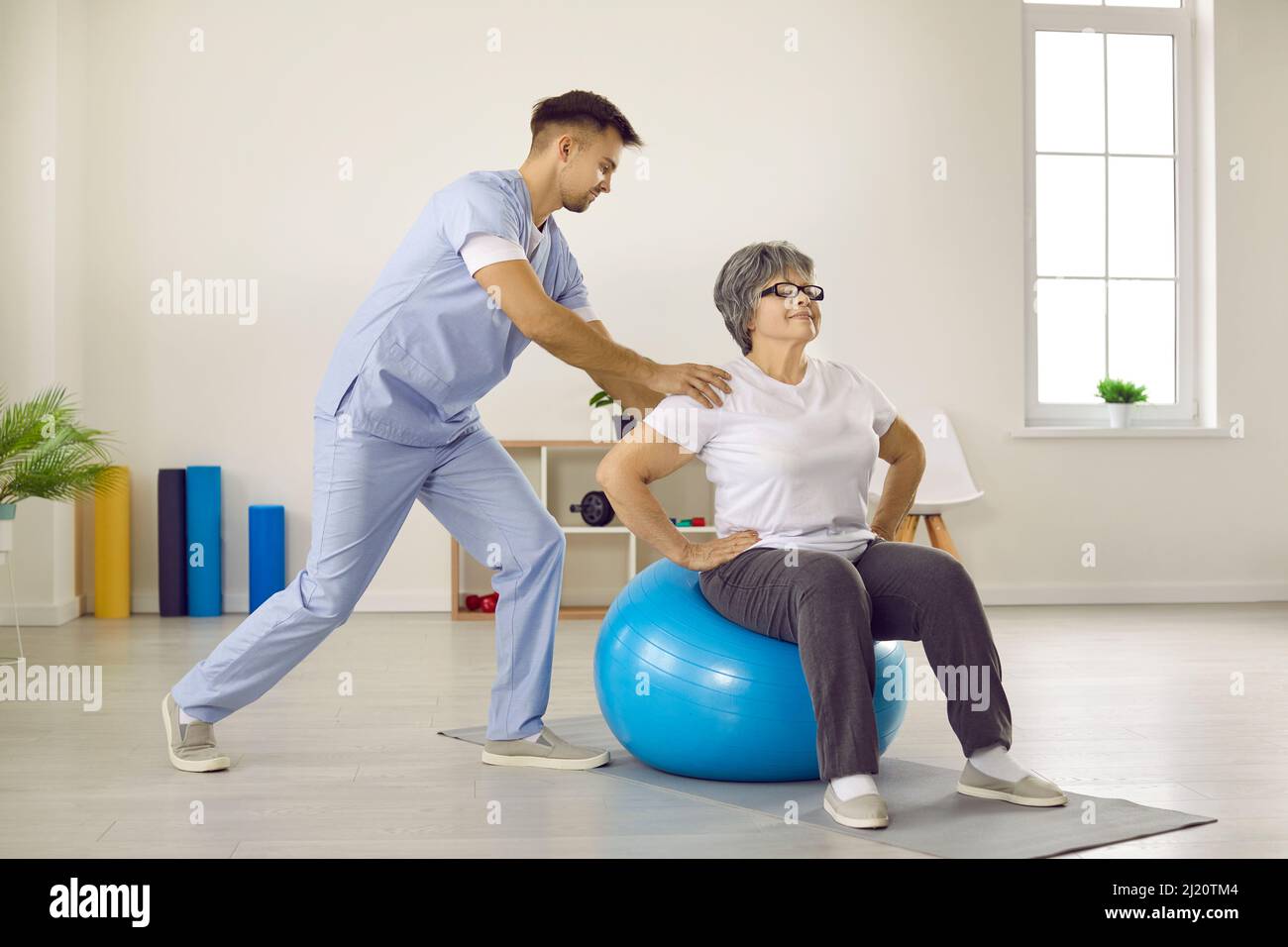 Male doctor with help of fitball trains elderly female patient in rehabilitation clinic. Stock Photo