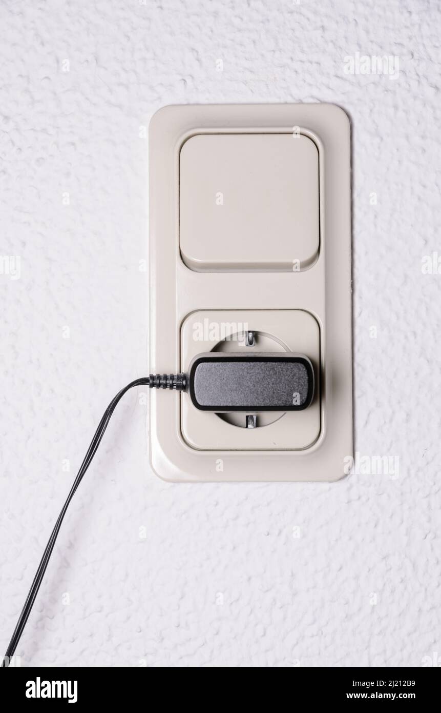 Modern light switch and german power outlet or socket on white wall indoors with black phone charger plugged in, Germany, Europe Stock Photo