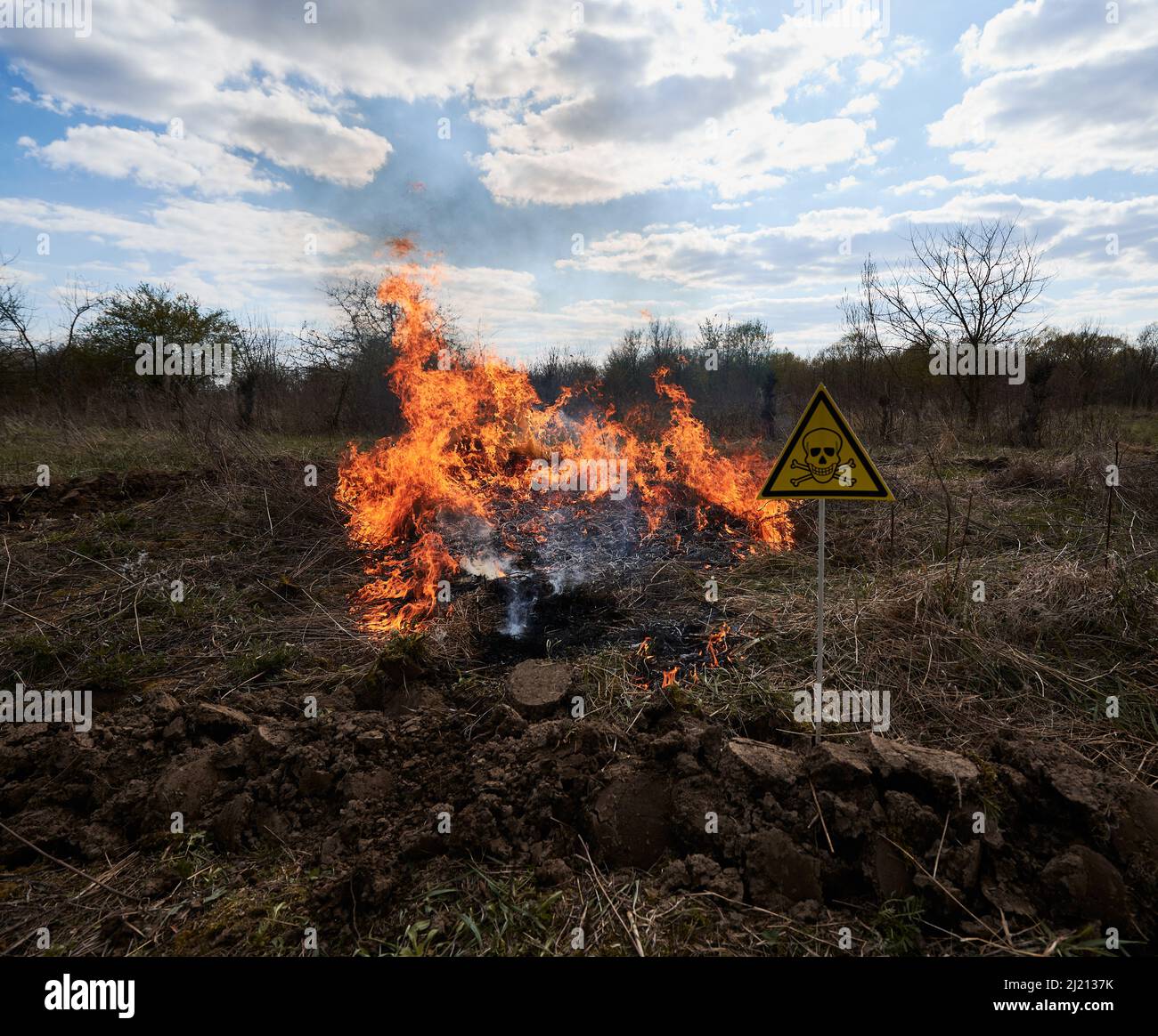 Burning dry grass and poison toxic sign under cloudy sky. Yellow triangle with skull and crossbones sign warning about danger in field with fire. Ecology, hazard and natural disaster concept. Stock Photo