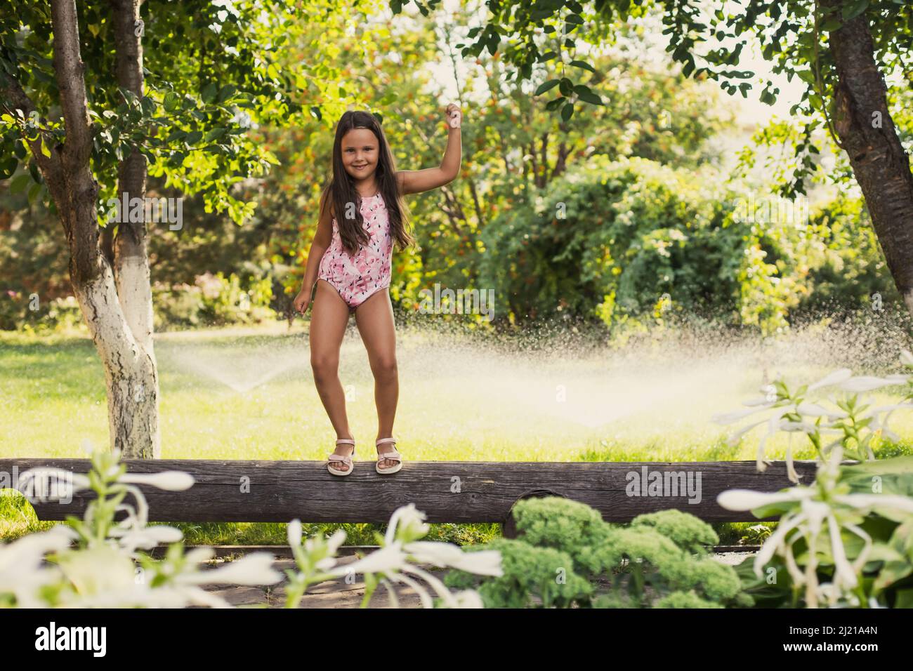 Shining young girl standing on log dancing indulging looking at camera with water sprinklers, house and green trees in background in daytime. Dreaming Stock Photo