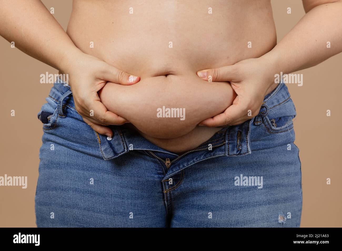 Caucasian obese Woman touching and showing big tummy wearing blue unzipped jeans on beige background. Sudden weight gain. Visceral fat. Body positive Stock Photo