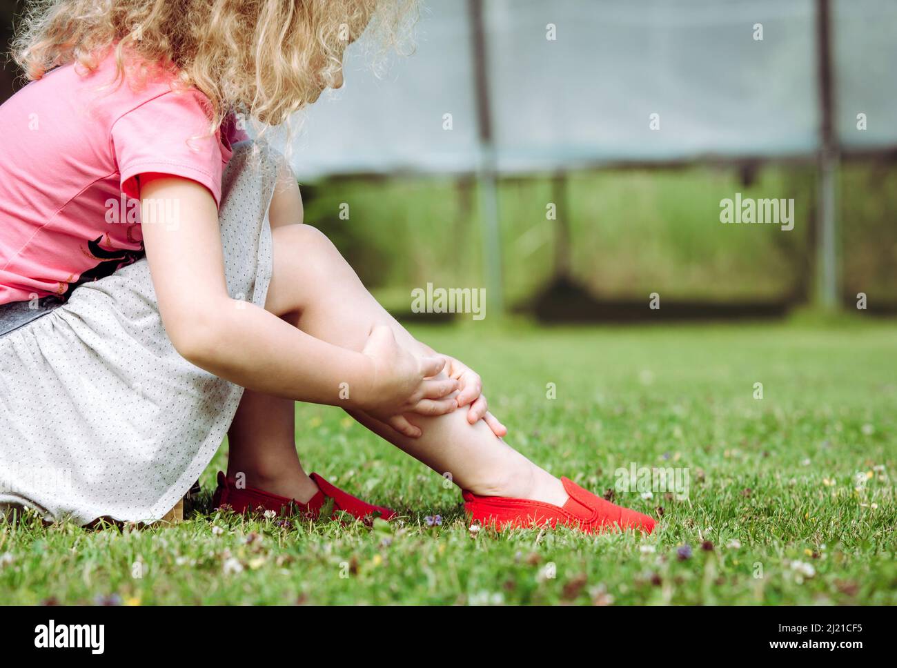 Trampoline jumping accident concept. Girl child holding sprained ankle with hands outdoors by the home garden trampoline. Hazards at home concept. Stock Photo