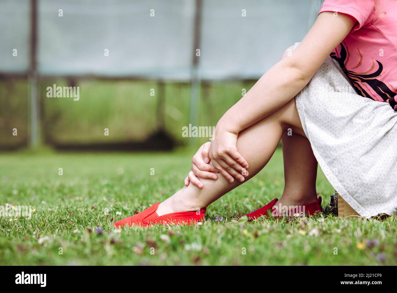 Trampoline jumping accident concept. Girl child holding sprained ankle with hands outdoors by the home garden trampoline. Hazards at home concept. Stock Photo