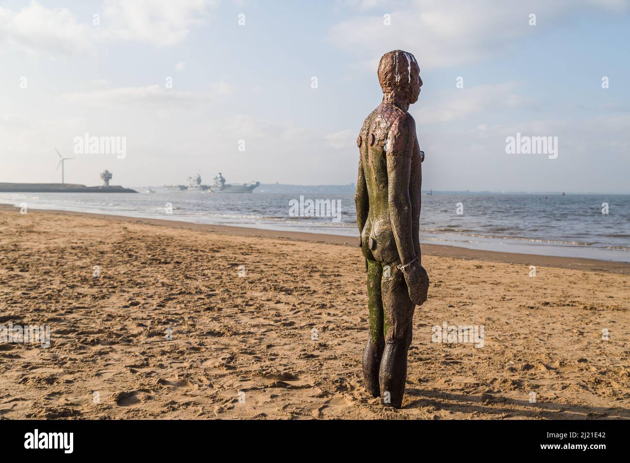 An Iron Man in the foreground waits patiently for aircraft carrier HMS Queen Elizabeth to exit the River Mersey estuary on her journey back to see see Stock Photo