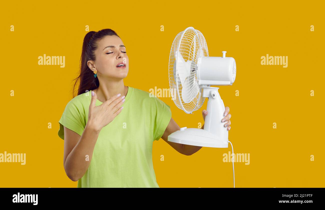 Exhausted young lady having headache because of summer heat, holding fan in her hand is exhausted from the heat, touching her forehead, yellow studio Stock Photo