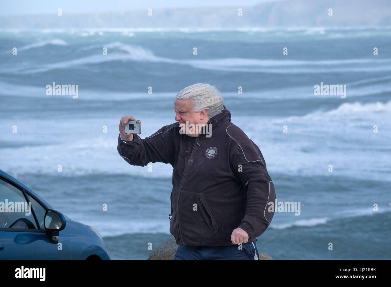 A man grimacing and using a mobile phone to record the wild weather brought by Storm Eunice in Newquay in Cornwall. Stock Photo