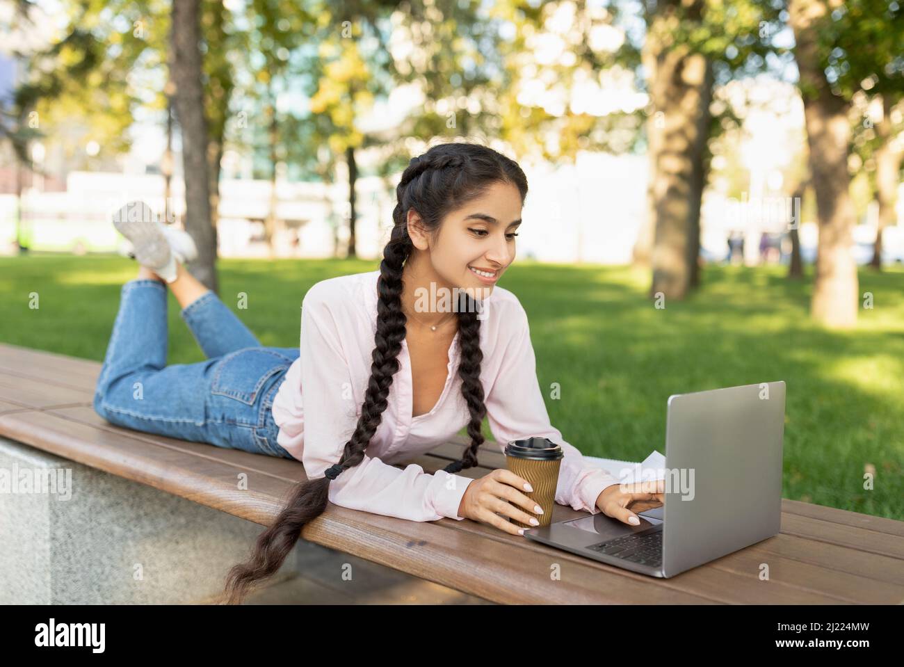 Rest at campus. Happy indian college student girl relaxing outdoors with coffee and laptop, lying on bench in park Stock Photo