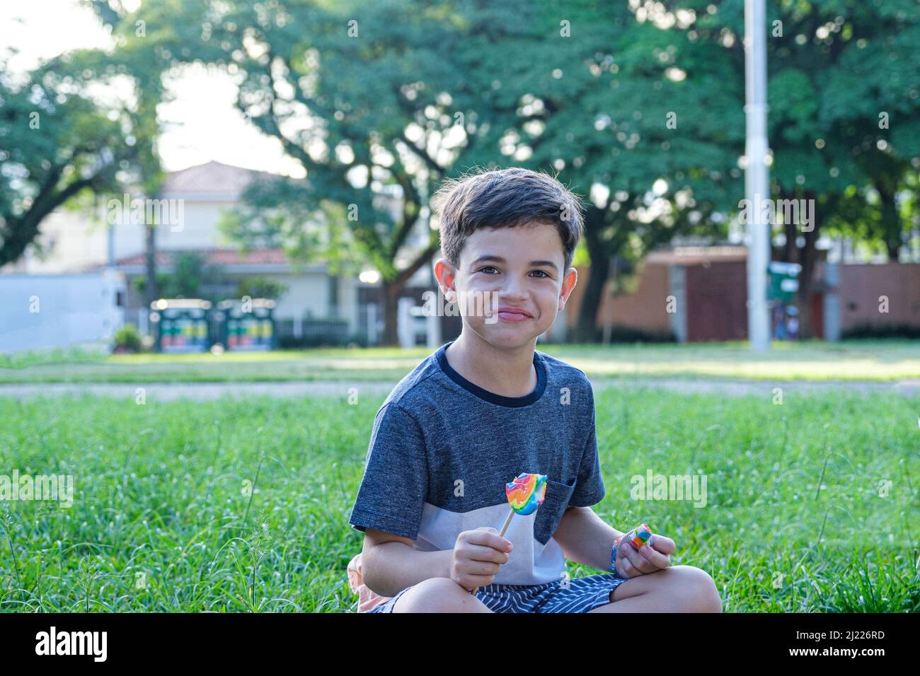 8 year old boy sitting on the lawn on a sunny afternoon, smiling and holding a lollipop. Stock Photo