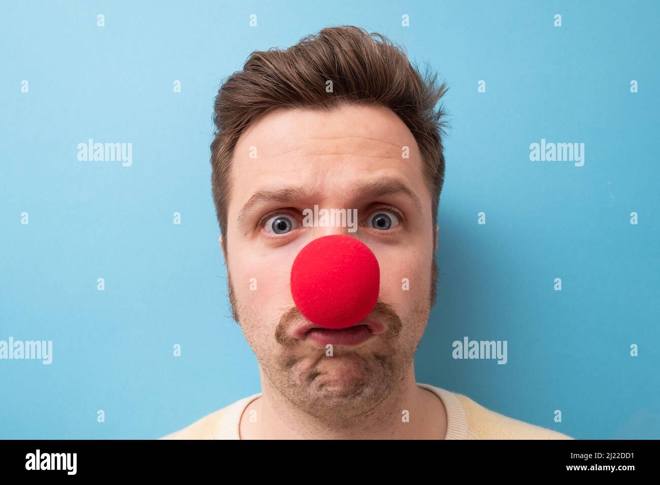Caucasian man wearing clown red nose isolated on blue background. He is upset that nobody came to his party. Stock Photo