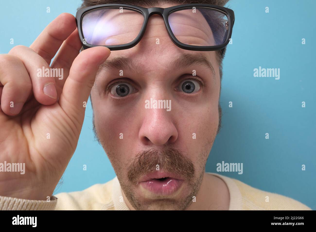 Young man with funny mustache in big glasses having doubts and with confuse face expression on isolated blue background Stock Photo
