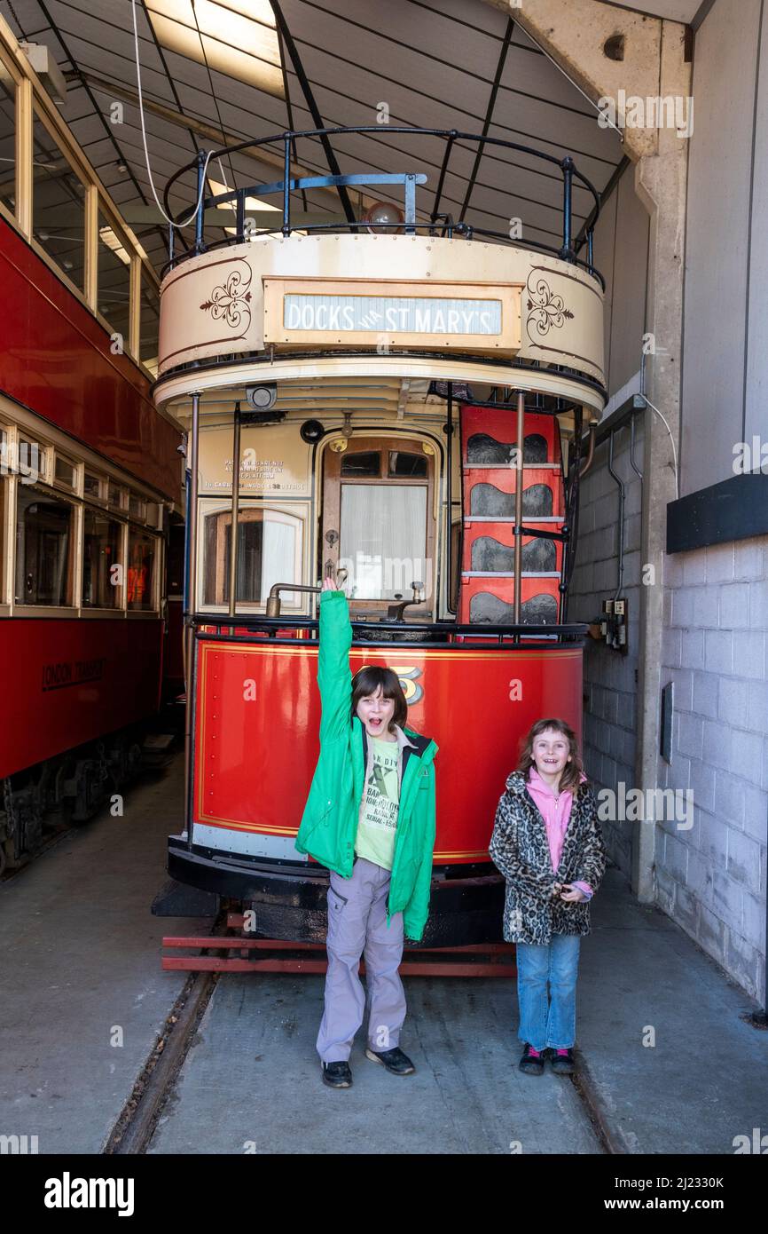 Derbyshire, UK – 5 April 2018: Two children pose in front of a vintage tram stored in the vehicle garage at Crich Tramway Village Stock Photo