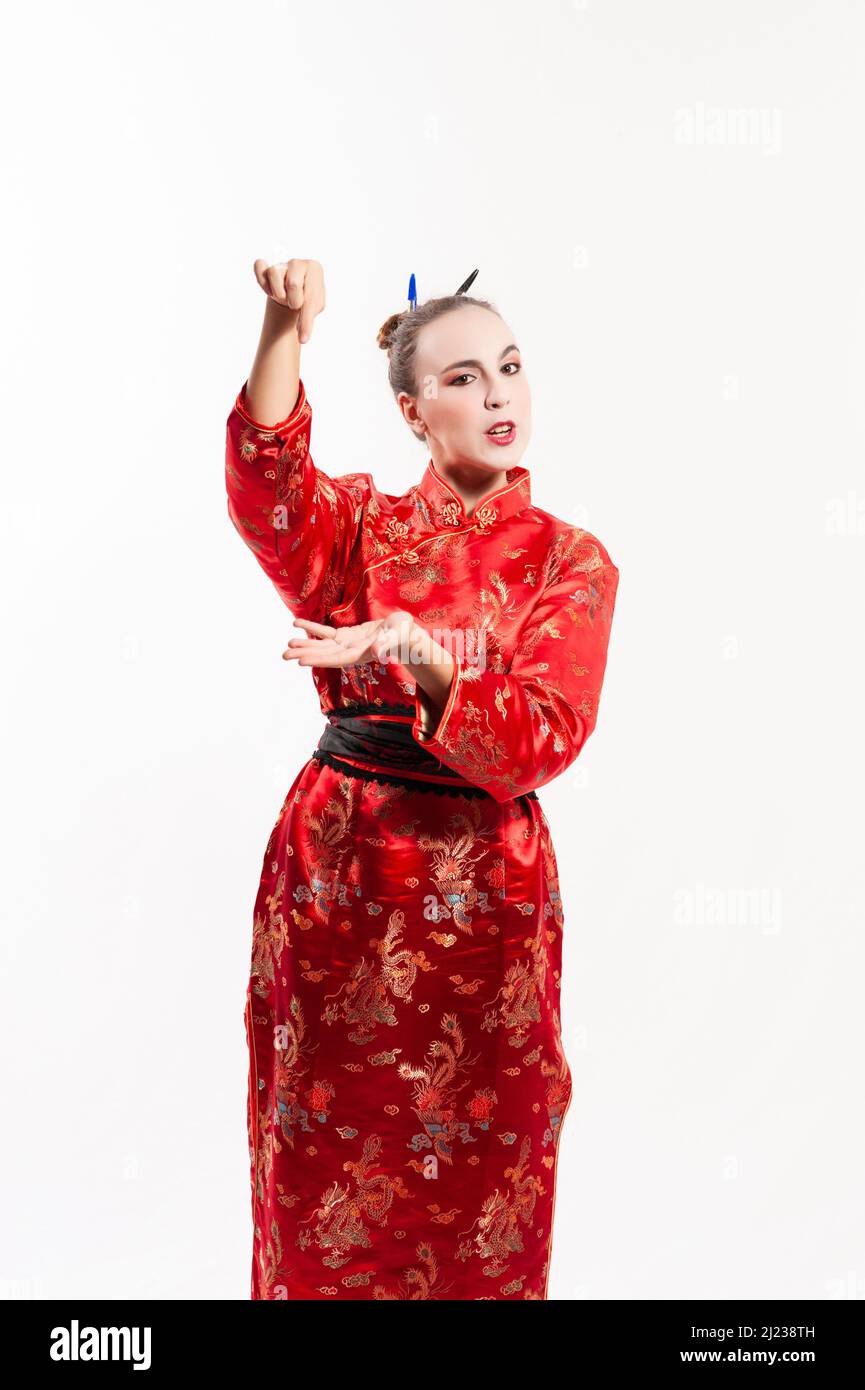 Young woman dressed in a traditional red Chinese costume, wearing white makeup, with a bun in her hair held in place with pens, raising her arm holdin Stock Photo