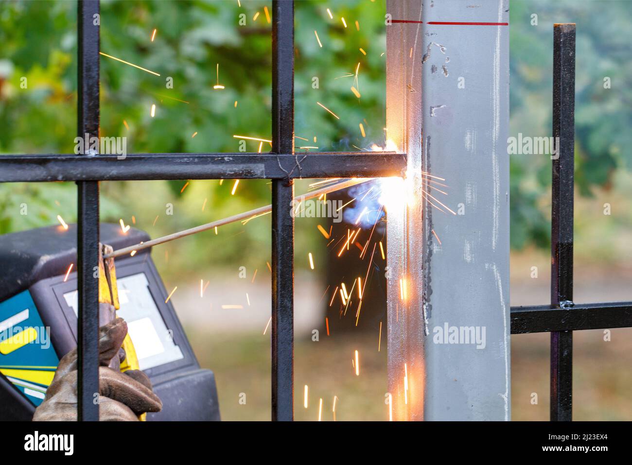 Bright sparks from the arc of electric welding fly in different directions against the background of a metal fence and a working welder in blur. Stock Photo