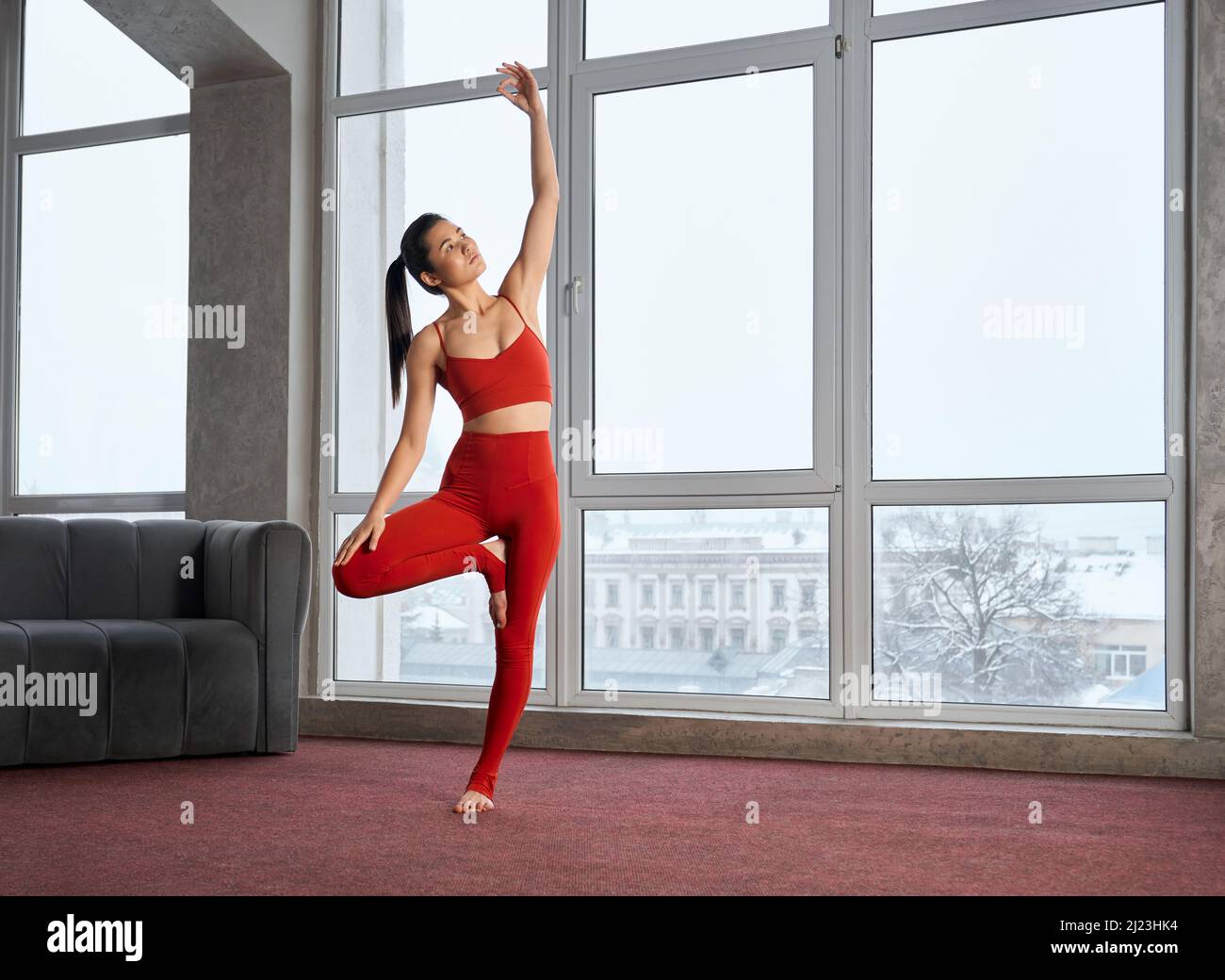 Side view of slim girl standing on one leg with raised hand indoors. Brunette female with ponytail practicing yoga, standing with bent leg, looking up. Concept of inner harmony. Stock Photo