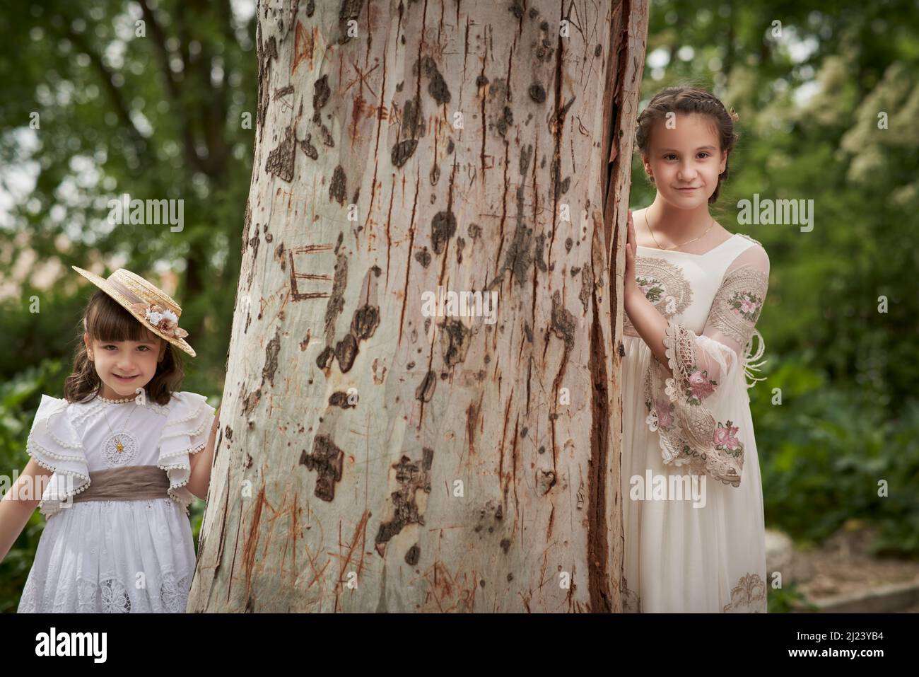 Communion girl posing with her sister next to a large tree Stock Photo