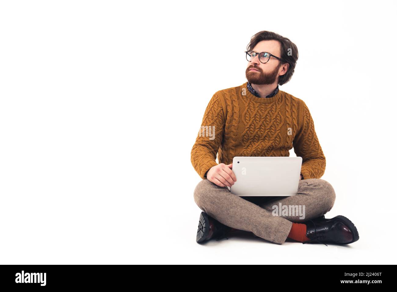 Full shot young attractive Caucasian man with beard and glasses sitting on the floor, legs crossed, keeping laptop lap, looking up away remote job concept isolated white background copy space . High quality photo Stock Photo