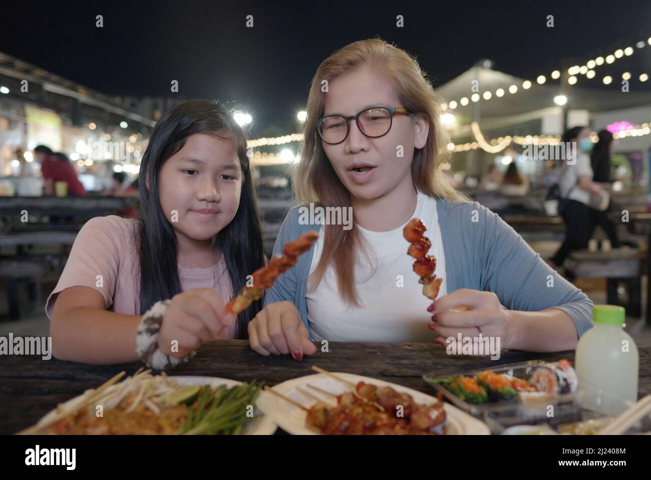 Mother and daughter eating BBQ in night street market Stock Photo