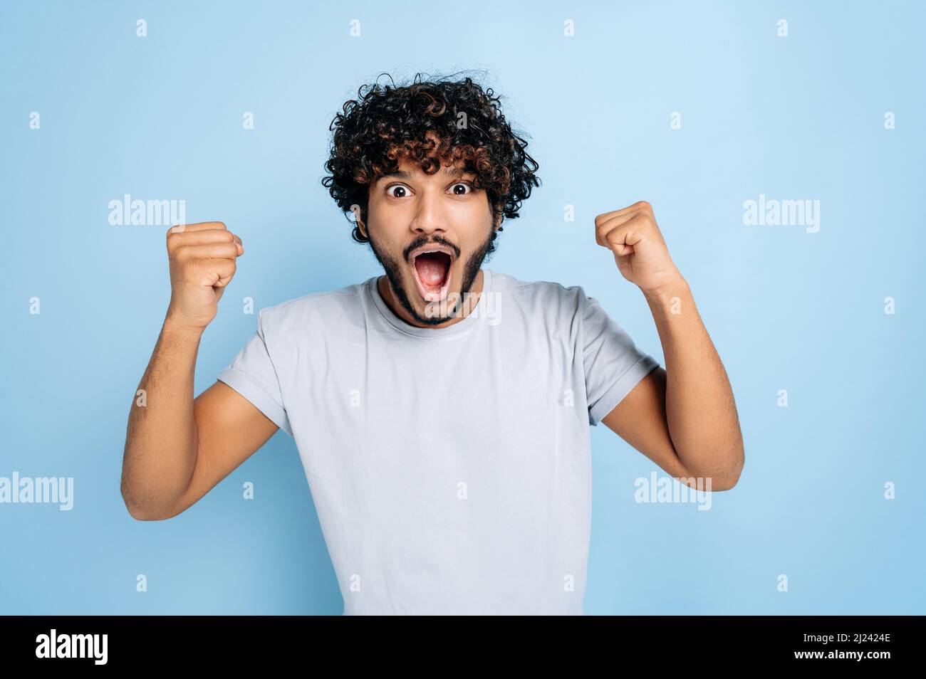 Enthusiastic amazed indian or arabic guy, in abasic t-shirt, rejoices in success, victory, win, gesturing with fists, looking at the camera, smiling, standing on an isolated blue background Stock Photo