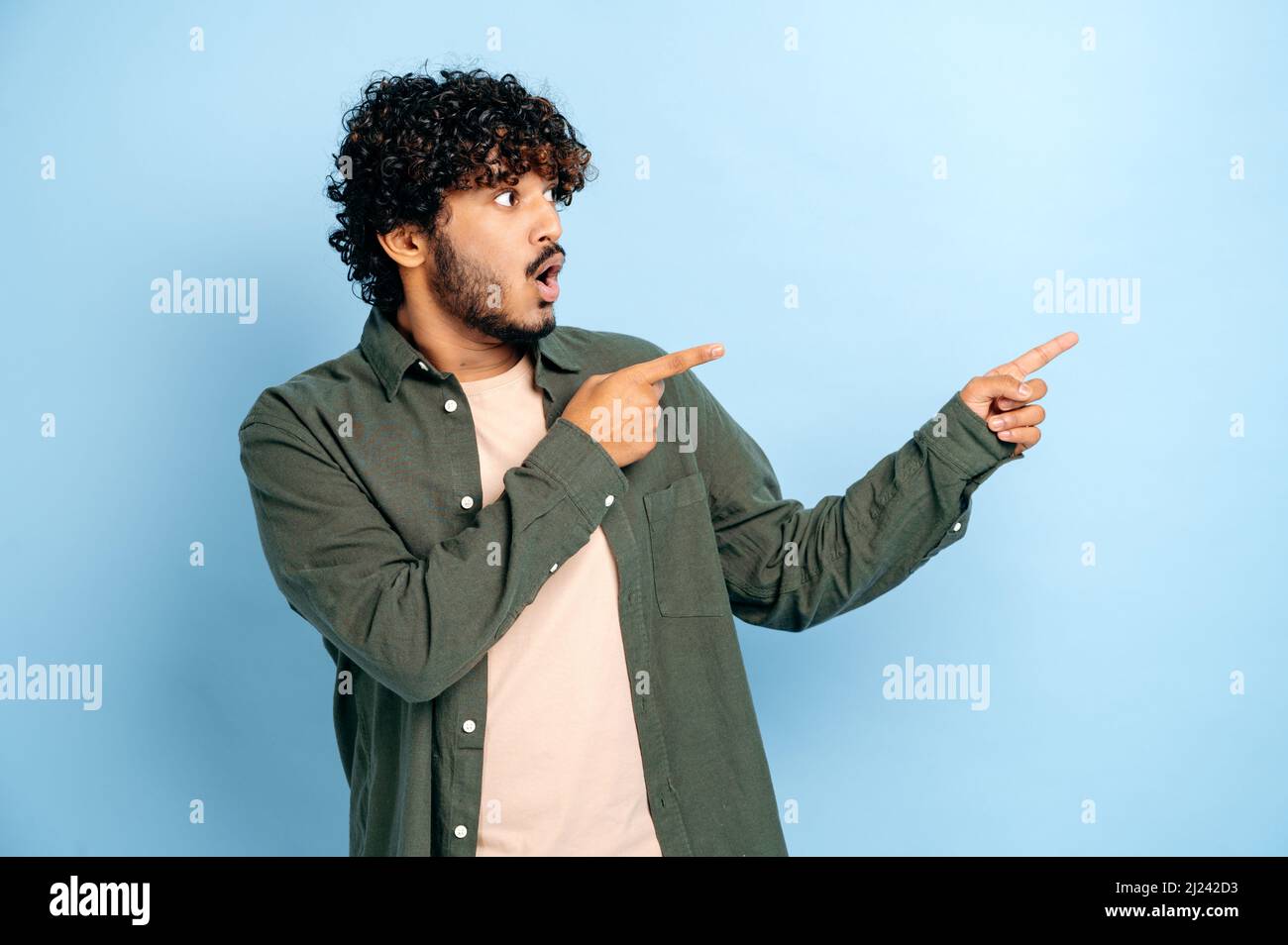 Shocked surprised indian or arabian curly haired guy in casual wear, points fingers to the side at mock-up empty space, stands on isolated blue color background, looks to the side, astonished Stock Photo