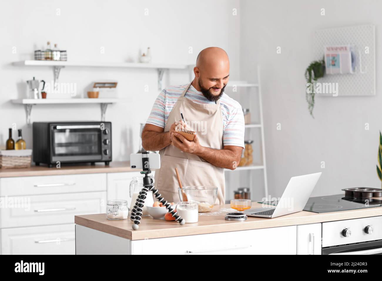 Handsome man making notes while watching cooking video tutorial on laptop in kitchen Stock Photo