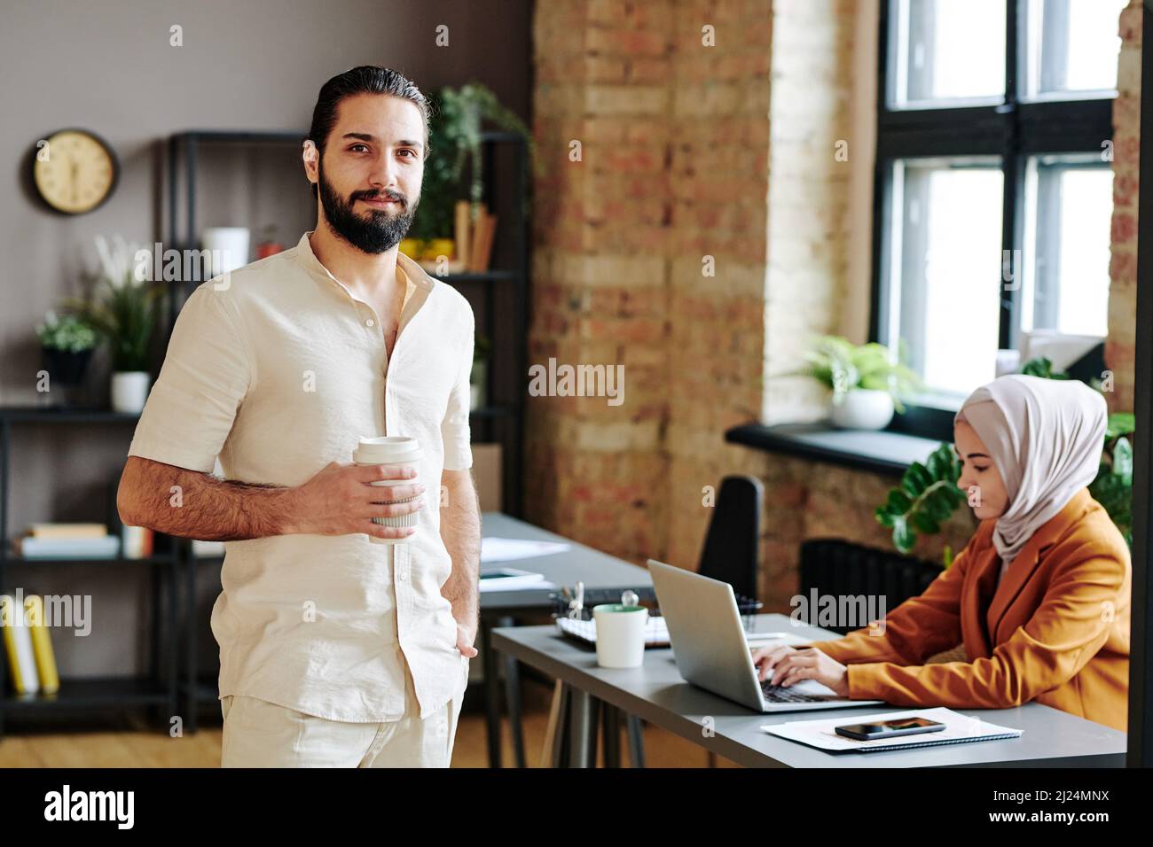 Happy young Muslim man in casual apparel holding cup of coffee in front of himself while standing against female colleague Stock Photo