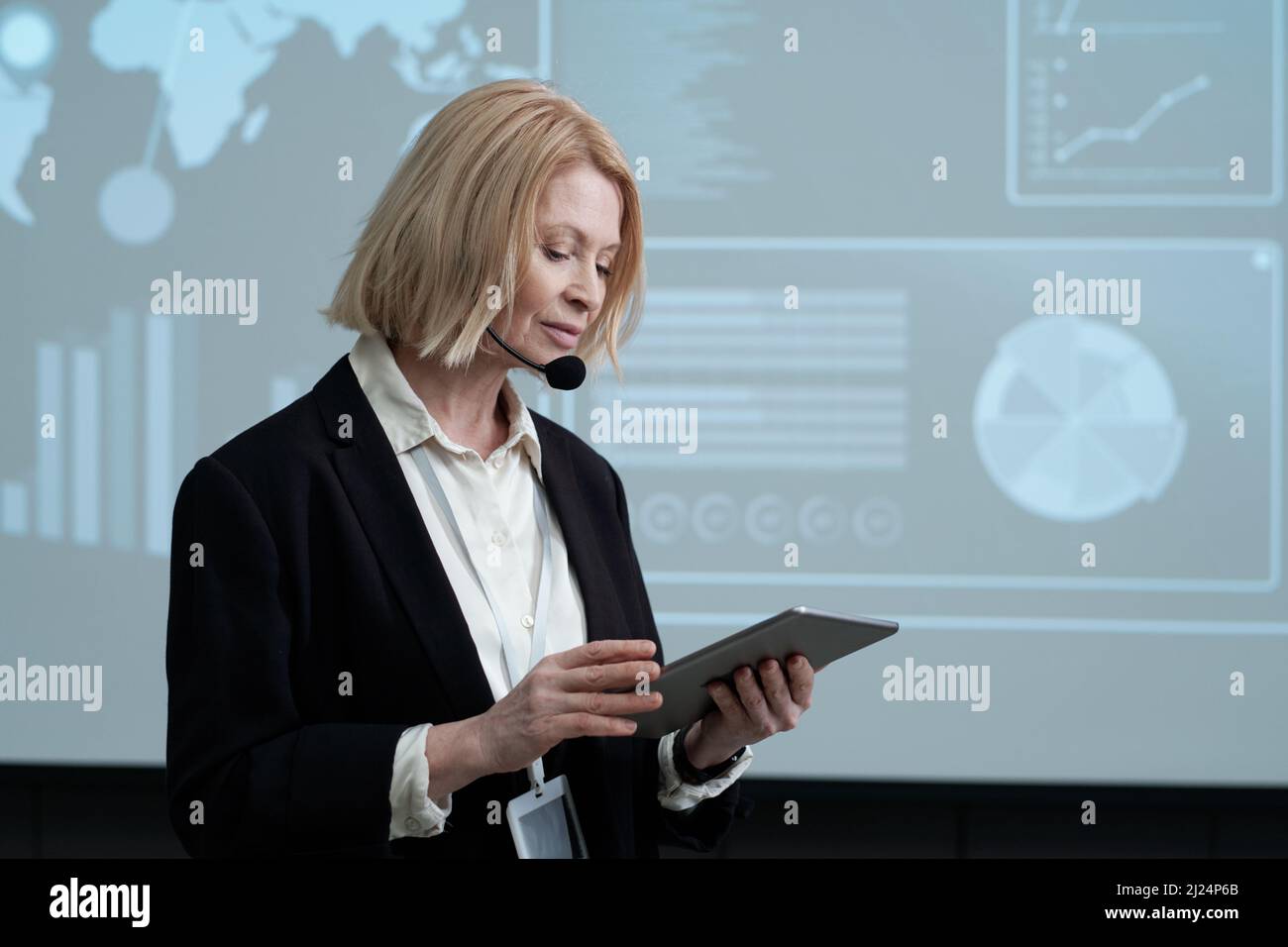 Serious female speaker or professor of economics with tablet reading lecture in auditorium while standing against interactive board Stock Photo