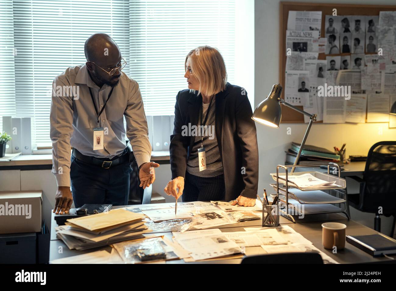 Two interracial agents of FBI discussing criminal profiles at meeting in bureau while one of them pointing at photo of suspect Stock Photo