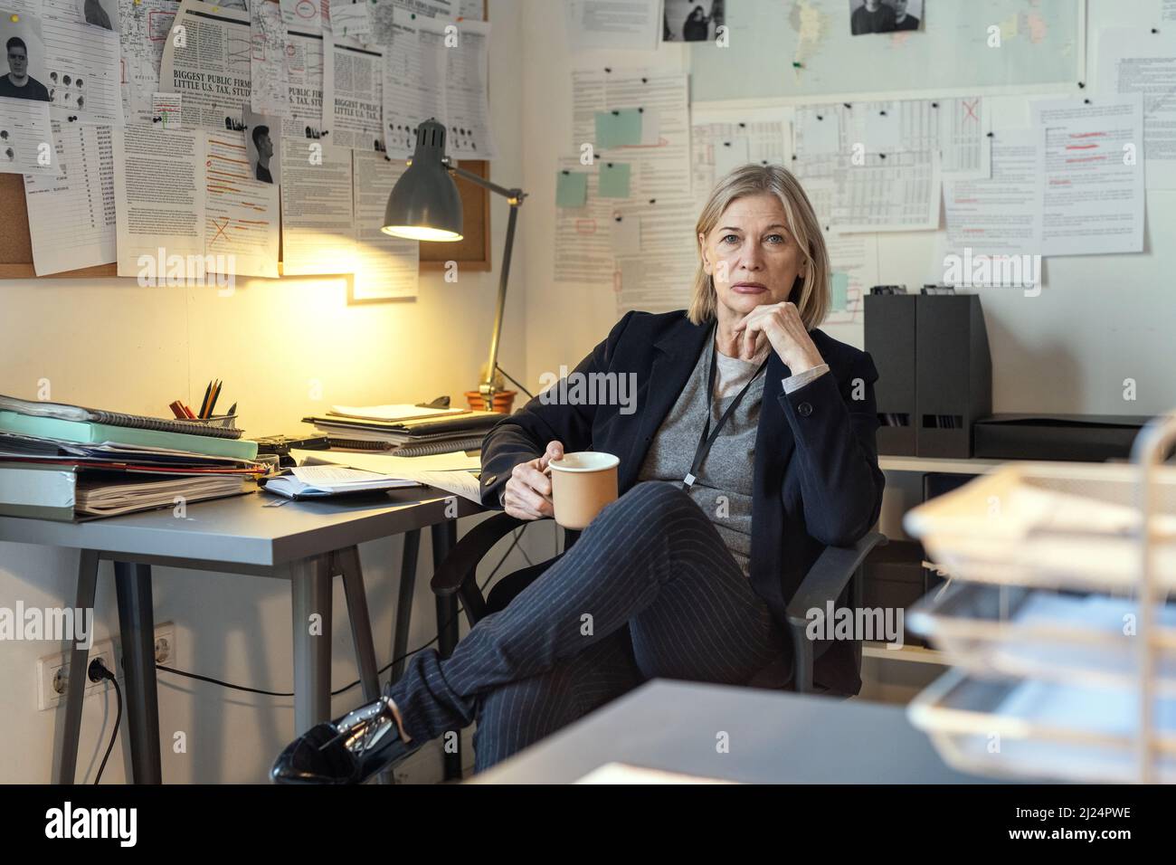Serious mature female detective with cup of coffee sitting by workplace against walls with criminal profiles and other working documents Stock Photo