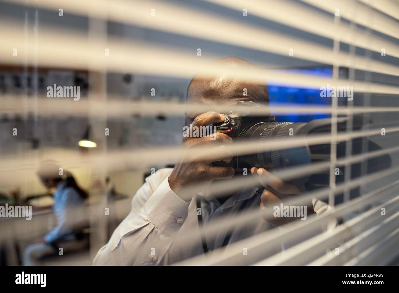 Young African American FBI agent or detective with photocamera taking photo through venetian blinds while standing by office window Stock Photo