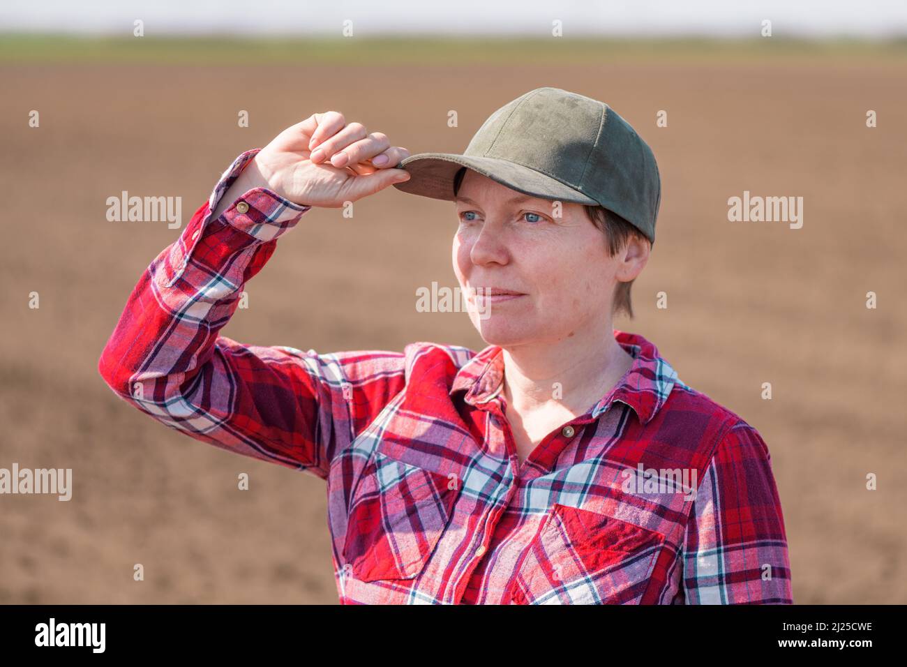 Portrait of female farmer with baseball cap and plaid shirt posing in plowed agricultural field prepared for seeding season, selective focus Stock Photo