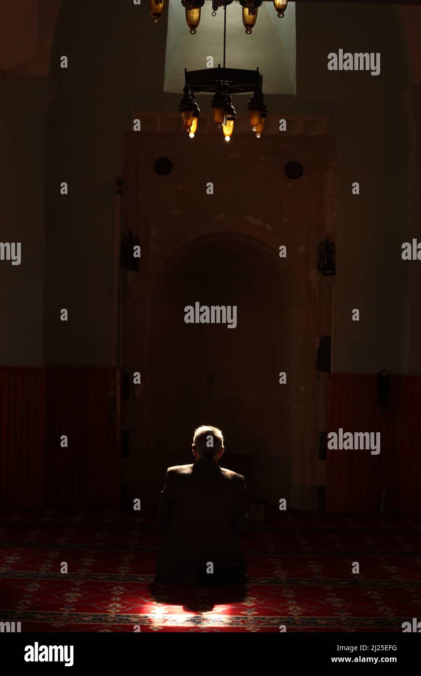 Back of unrecognizable white old man praying in dark mosque with a beam of sunlight falling over head and shoulders. Prayer area with red rug, carpet Stock Photo
