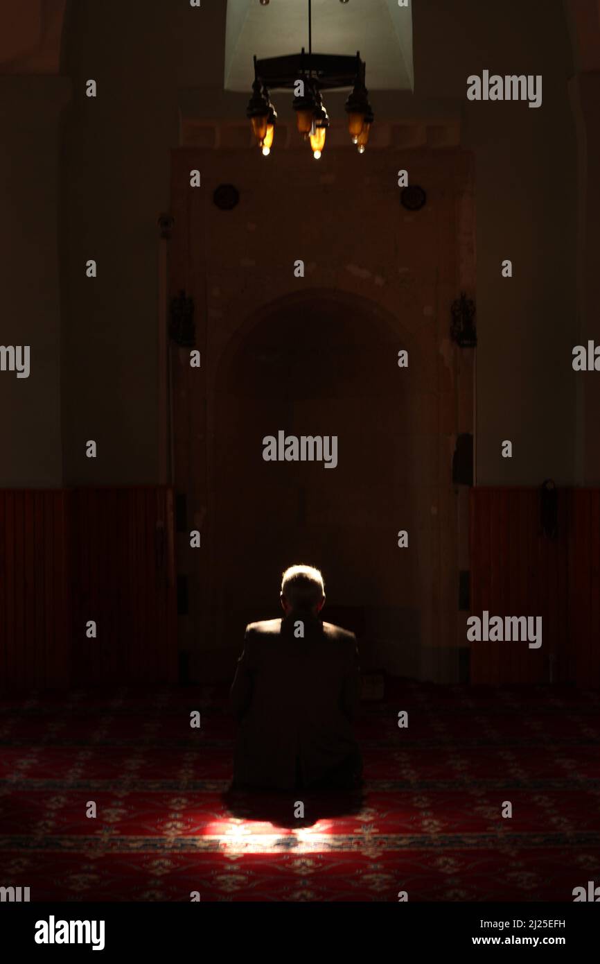 Back of unrecognizable white old man praying in dark mosque with a beam of sunlight falling over head and shoulders. Prayer area with red rug, carpet Stock Photo