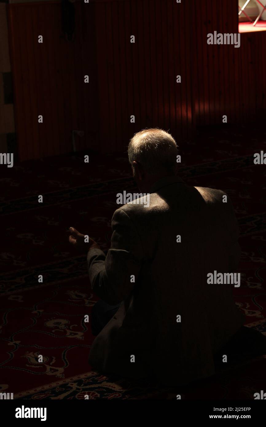 Back of unrecognizable white old man praying in dark mosque with a beam of sunlight falling over head and shoulders. Prayer area with red rug, carpet Stock Photo