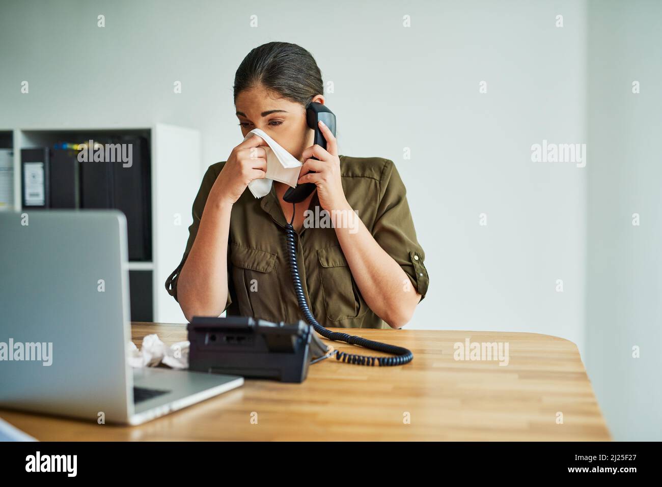 Its an unbearable condition to work in. Shot of a young businesswoman blowing her nose while speaking on a phone in an office. Stock Photo