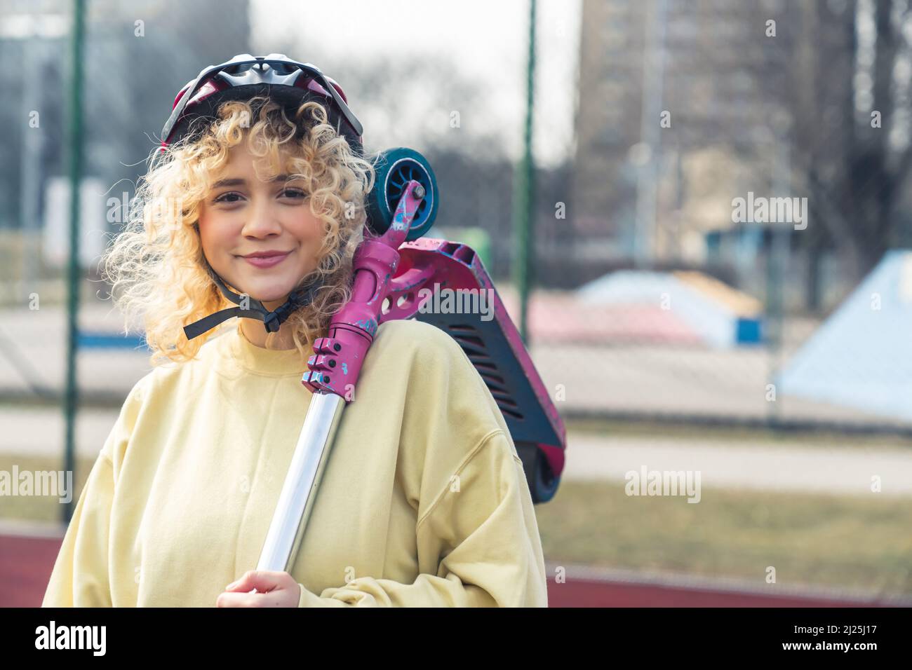 Pretty blond curly haired Caucasian woman holds a pink scooter on her shoulder, wears yellow sweater and helmet, standing outside on the sports field portrait copy space . High quality photo Stock Photo