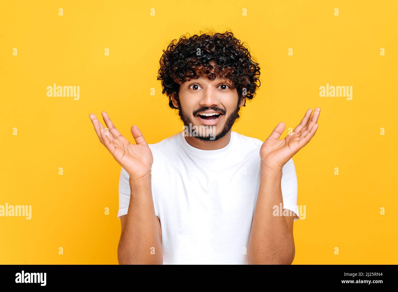 Surprised confused indian or arabian guy with curly hair, wearing white t-shirt, looking at camera in disbelief, spreading his arms to the sides, shrugging, standing over isolated orange background Stock Photo