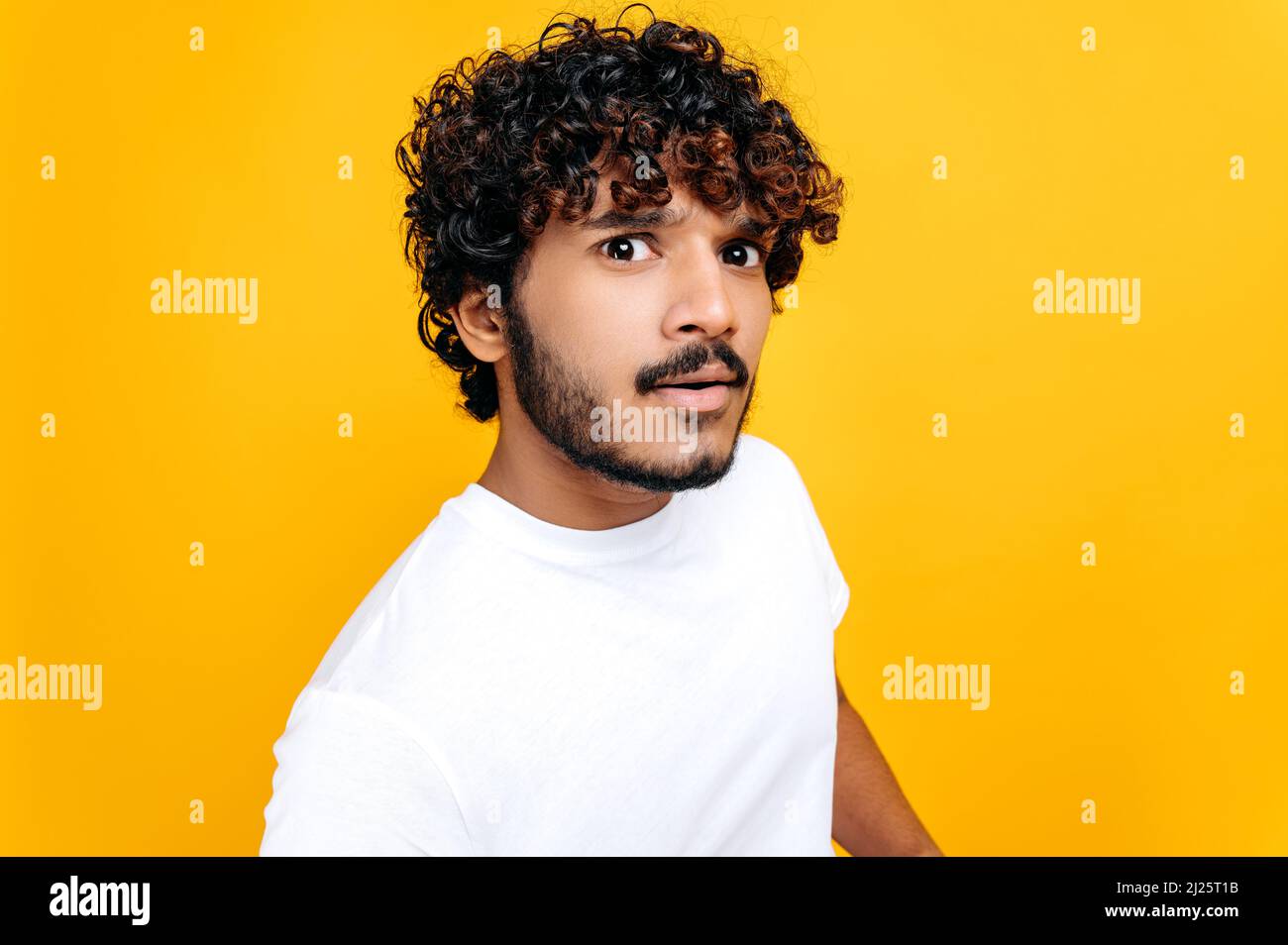 Close up of confused puzzled curly haired indian or arabian guy in white t-shirt, looking questioningly at the camera, while standing over isolated orange background Stock Photo