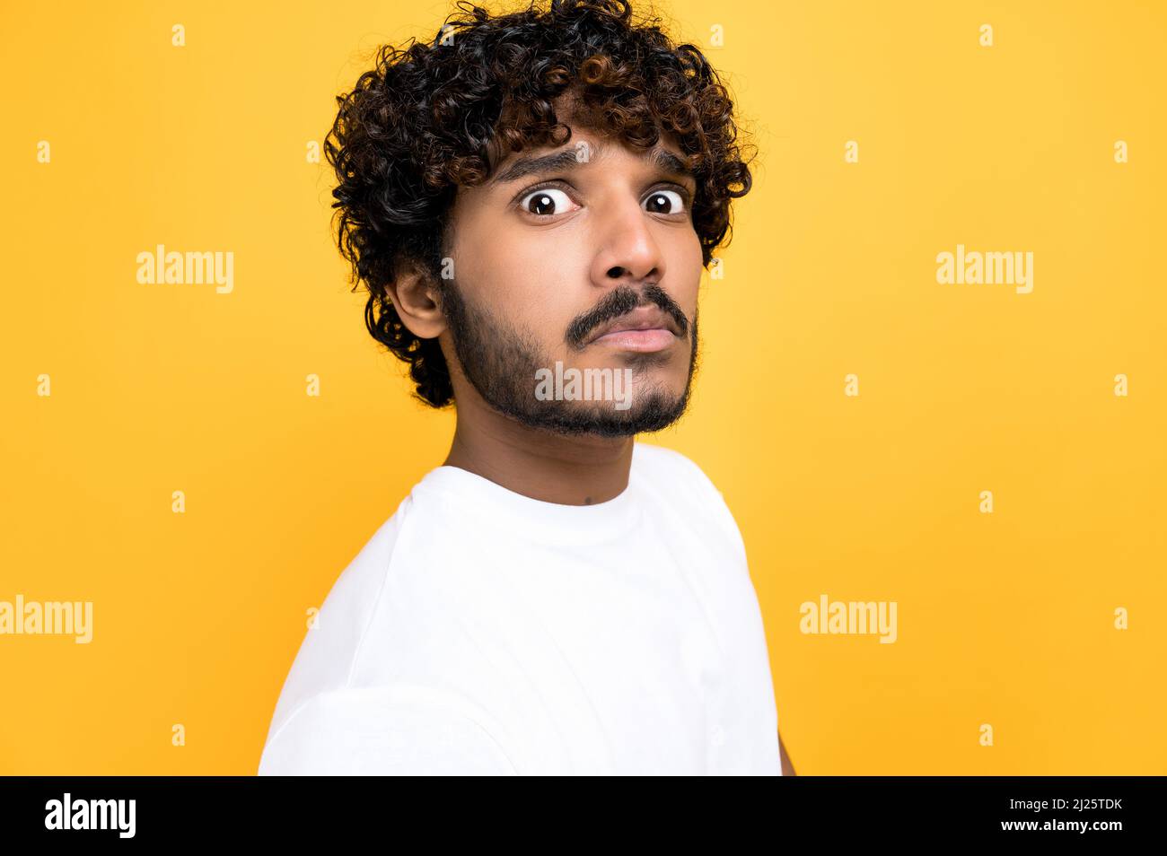 Close-up photo of a scared confused indian or arabian curly haired guy,  looking at camera in disbelief, standing over isolated orange background  wearing white t-shirt Stock Photo - Alamy