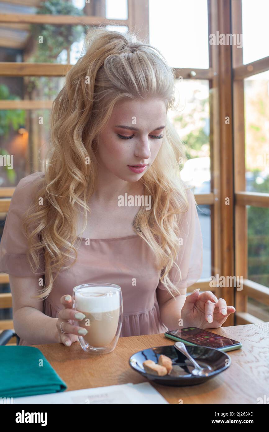 Young pretty woman holding coffee cup and looking at smartphone while sitting at cafeteria. Stock Photo